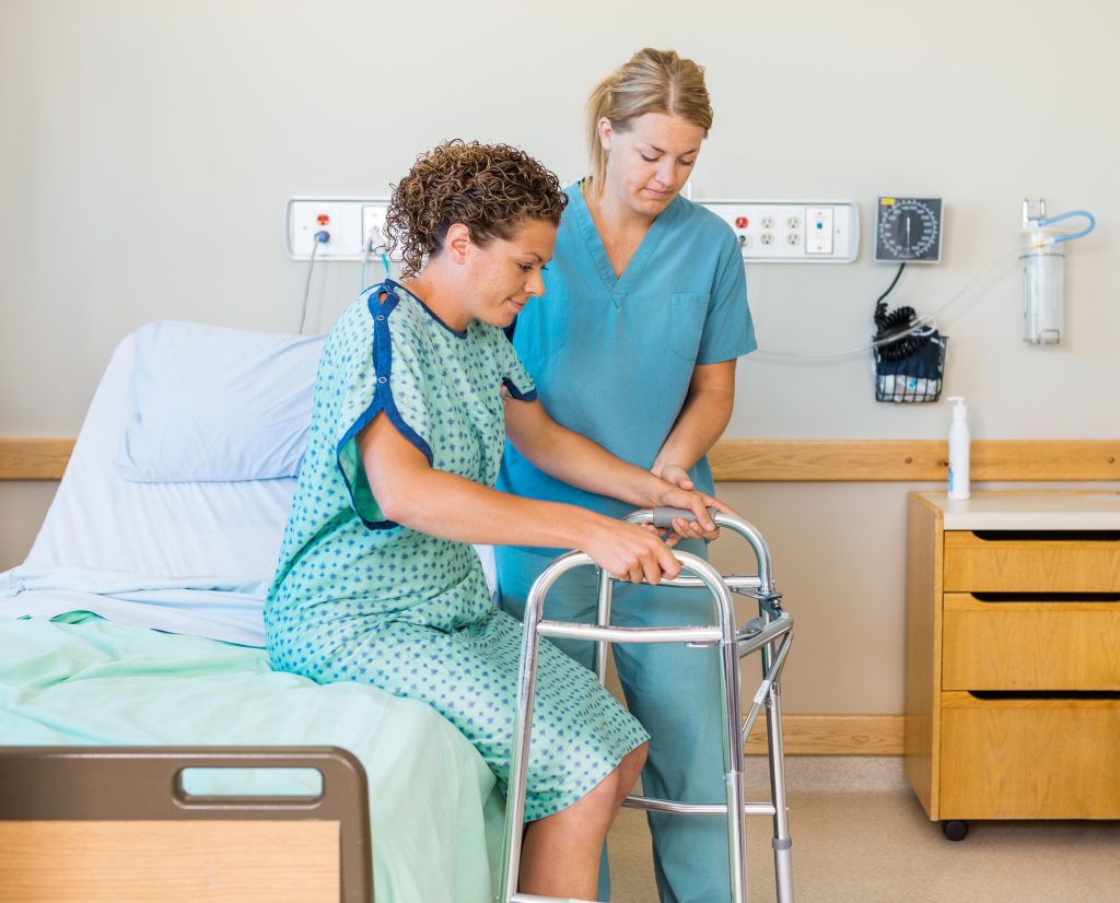 Caucasian woman sitting on a hospital bed holding a folding rolling walker while a female caucasian nurse, physical therapist, or occupational therapist prepares to help on the side. Acute Care Facility