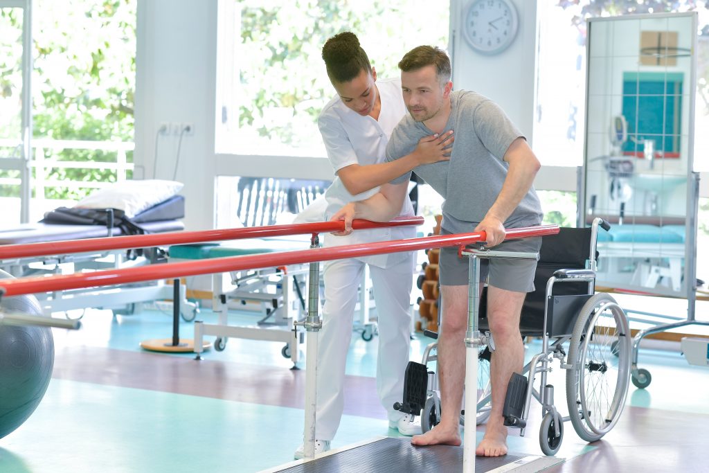 African American female healthcare worker,: physical therapist. Assisting a patient with gait training in the parallel bars. Inpatient Rehabilitation Facility.