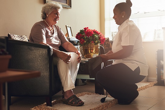 Caucasian older adult woman sitting on couch being cared for by an African American female nurse kneeling in front. Rehab for elderly after hospital stay.