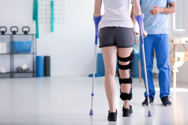 A picture of a woman in a right leg brace using forearm/lofstrand crutches and doctor/therapist standing in front. 