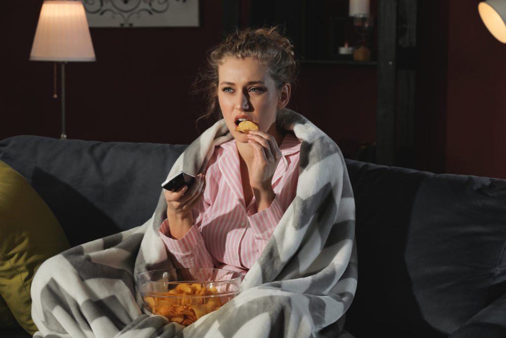 Caucasian woman sitting on couch late at night, eating potato chips