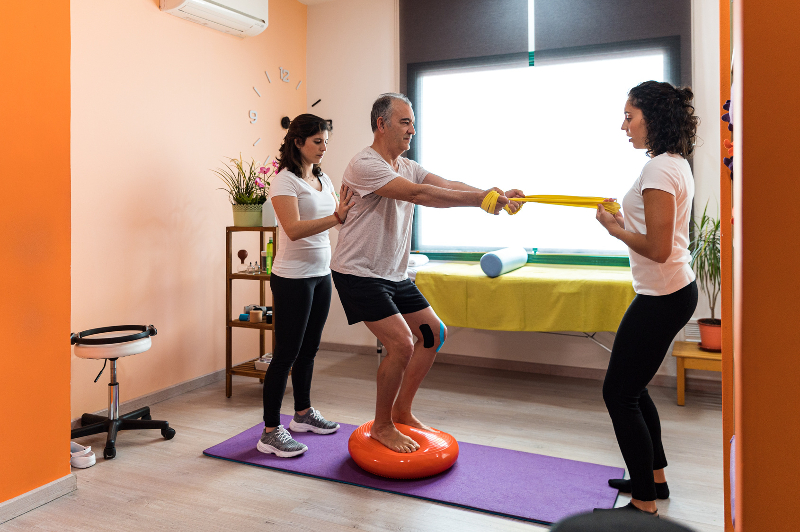 Caucasian man working on balance pad exercises with 2 female physiotherapist